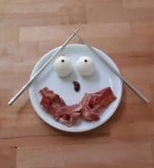 mit japanischen Eß-Stäbchen auf weißem Teller zu einem Gesicht dekorierte Eier, Schinken, Oliven, Pfefferkörner
